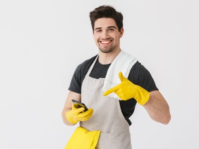 Handsome brunette houseman wearing apron standing isolated over white background, showing mobile phone